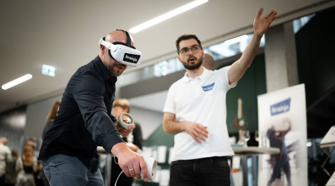 VR-Brille im Einsatz: Ein Mann testet die VR-Anwendung von brainjo und wird von einem Mitarbeiter eingewiesen.